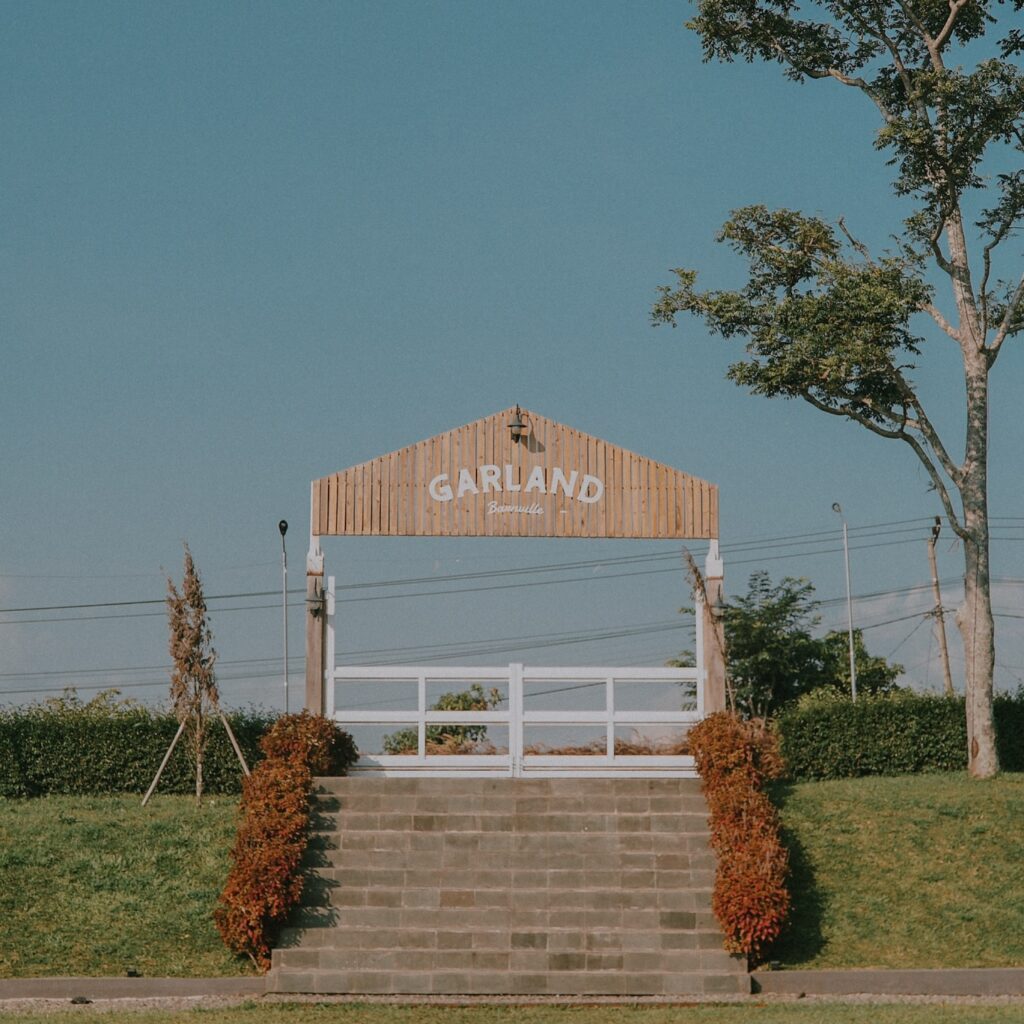 Gerbang masuk Garland Barnville dengan papan nama kayu bergaya rustic di bawah langit biru cerah, dikelilingi semak dedaunan coklat kemerahan dan rumput hijau yang vibrante
